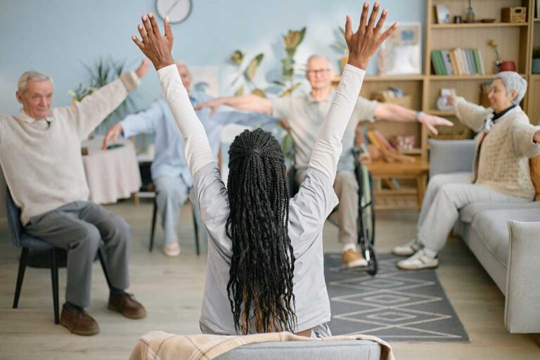 A group of older adults participating in a light chair exercise class in a bright, welcoming community room.