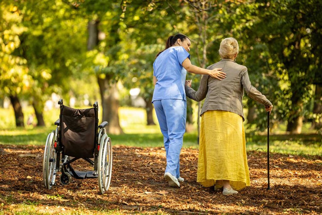 A caregiver assisting an older adult with medication at a small dining table in a bright, homey setting.